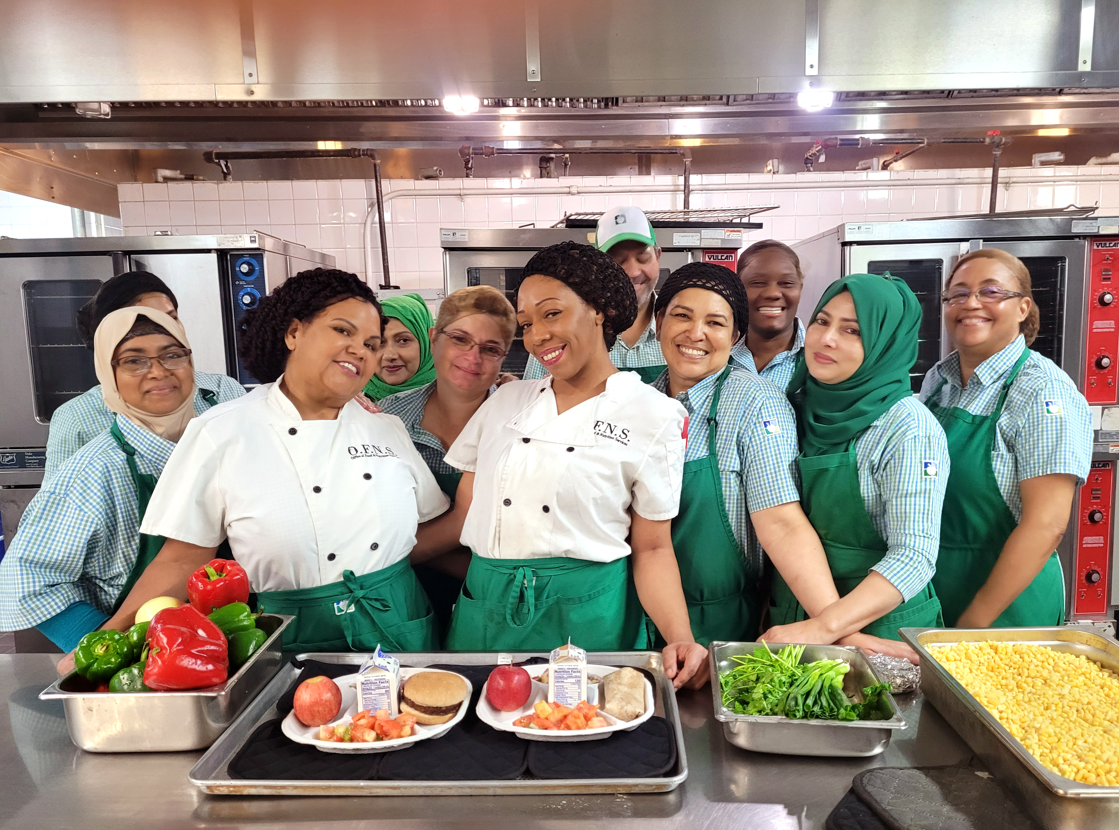 Senior Lunch Aide Shanna Brown (center) displays two vegetarian choices available for students. Local 372 members from left: Jesmin Akter, Tamanna Parvin, Second Cook Maria Deleon, Farzana Boby, Marilyn Ortiz, Shanna Brown, Carmen Morel, Samuel Cotto, Shanier Brown, Shahina Chowdhury, and Maria Diaz. Photo by Acacia Rodriguez